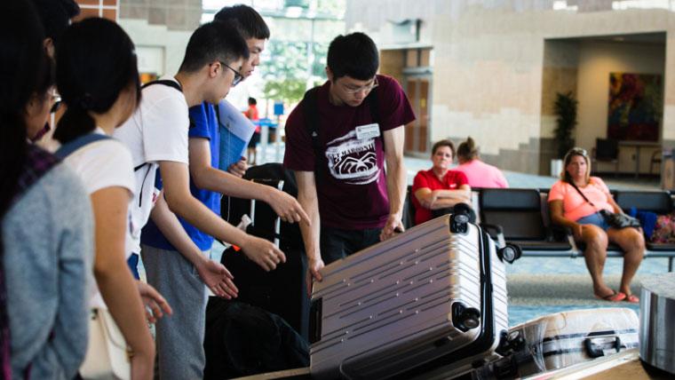 A group of young adults standing by an airport baggage carousel as one reaches for a silver suitcase