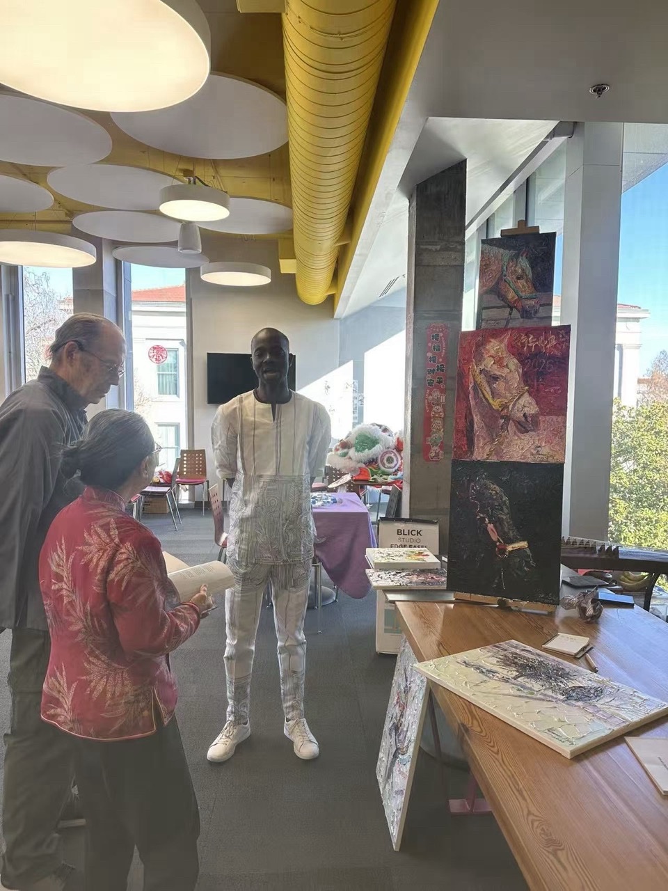 An art exhibition space where visitors look at displayed horse paintings while a man stands nearby in traditional patterned attire.