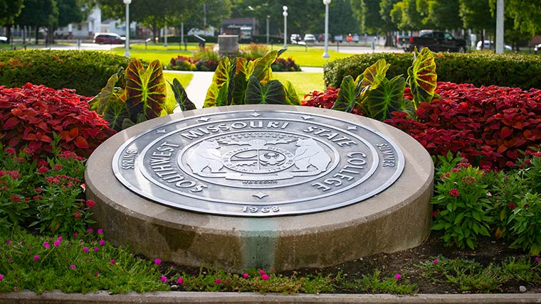 University seal in front of Carrington Hall, surrounded by colorful flowers and plants.