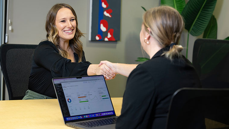 Two businesswomen shake hands at the efactory. A laptop sits between them.