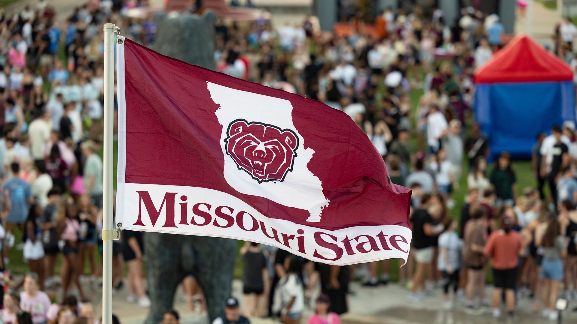 A Missouri State flags while a crowd of students attend an event in the background.