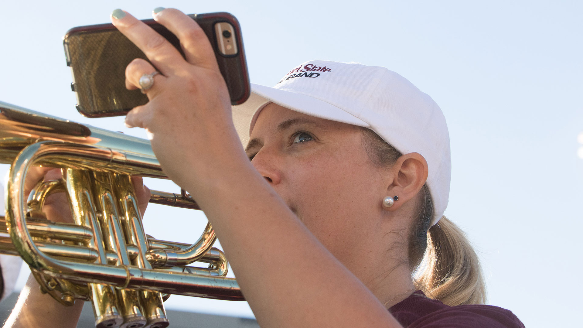 A student plays a trumpet while holding her phone up to take a picture.