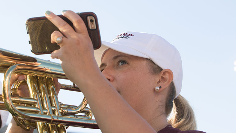 A student plays a trumpet while holding her phone up to take a picture.