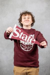 A male holding a maroon pendant that says "State" and smiling at the camera