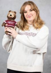 Image is of a female holding up a stuffed bear and smiling at the camera.