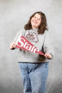 A female holding a maroon pendant that says "state" and smiling at the camera.