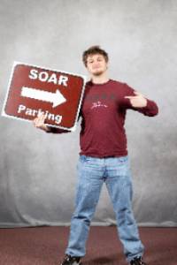 A male holding up a sign that says "SOAR" parking and smiling at the camera.
