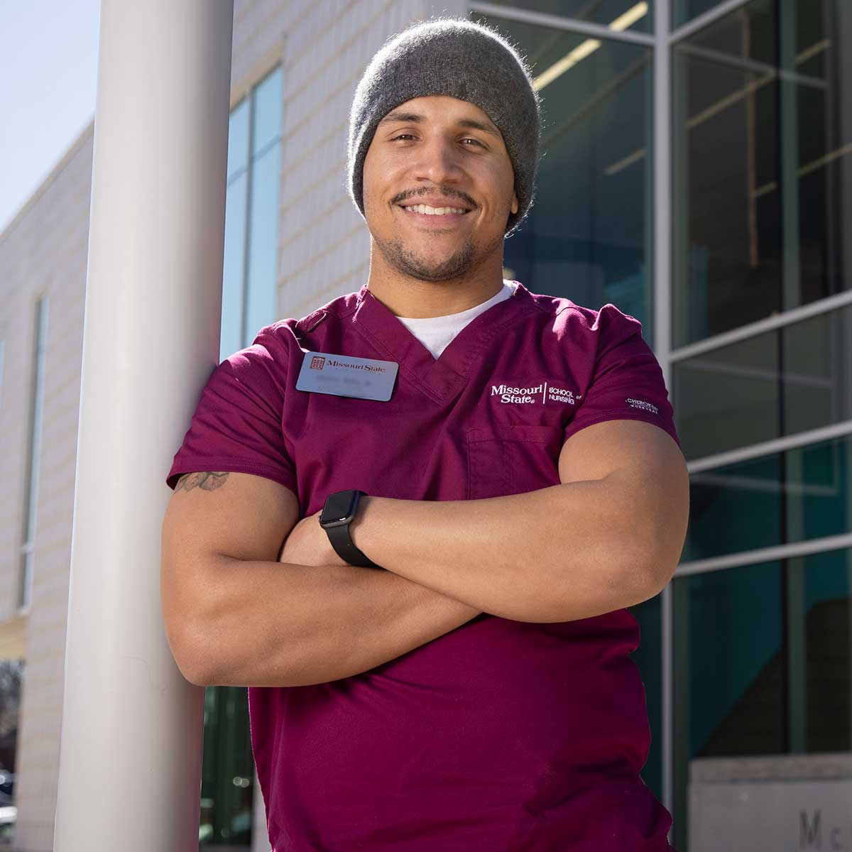A nursing student in scrubs poses outside of an academic building on campus.