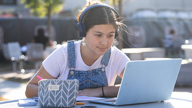 A student with headphones studying outside on her laptop on a nice, sunny day.