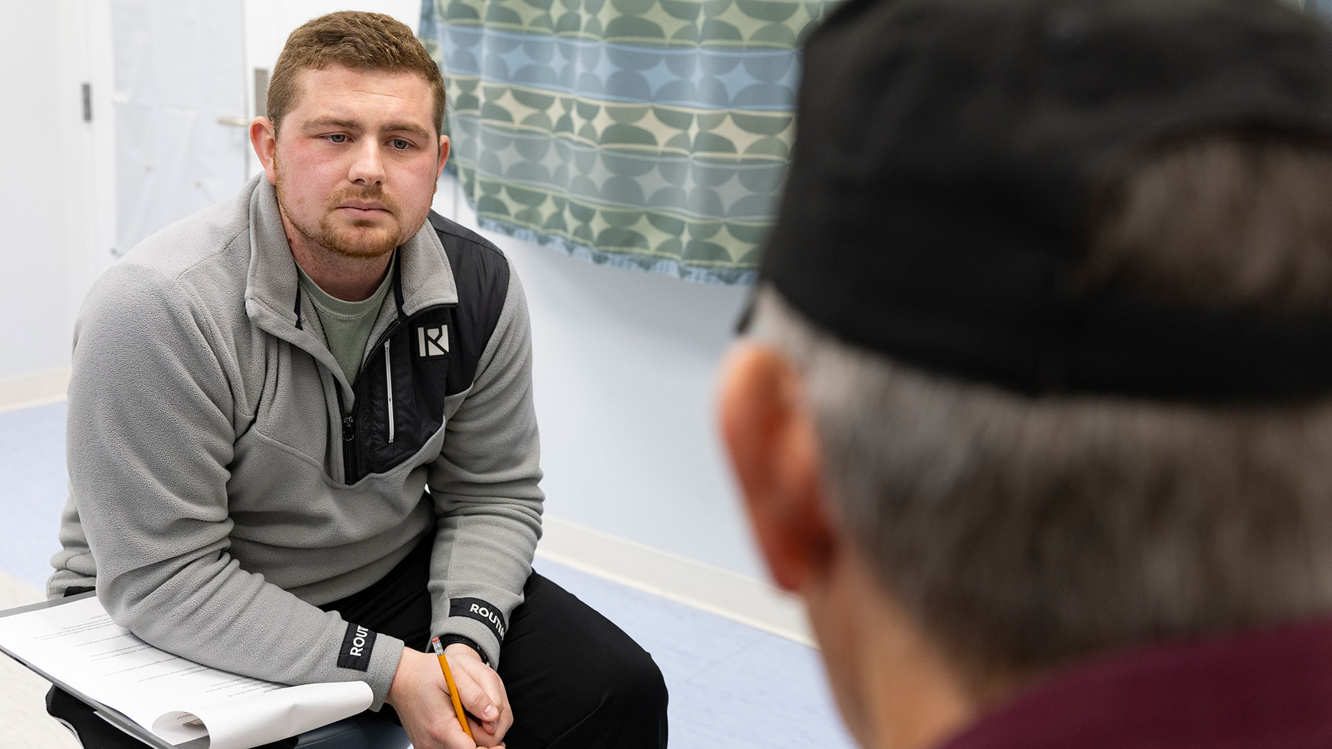 A graduate nursing student listens to his patient while holding onto a pencil and a pad of paper.
