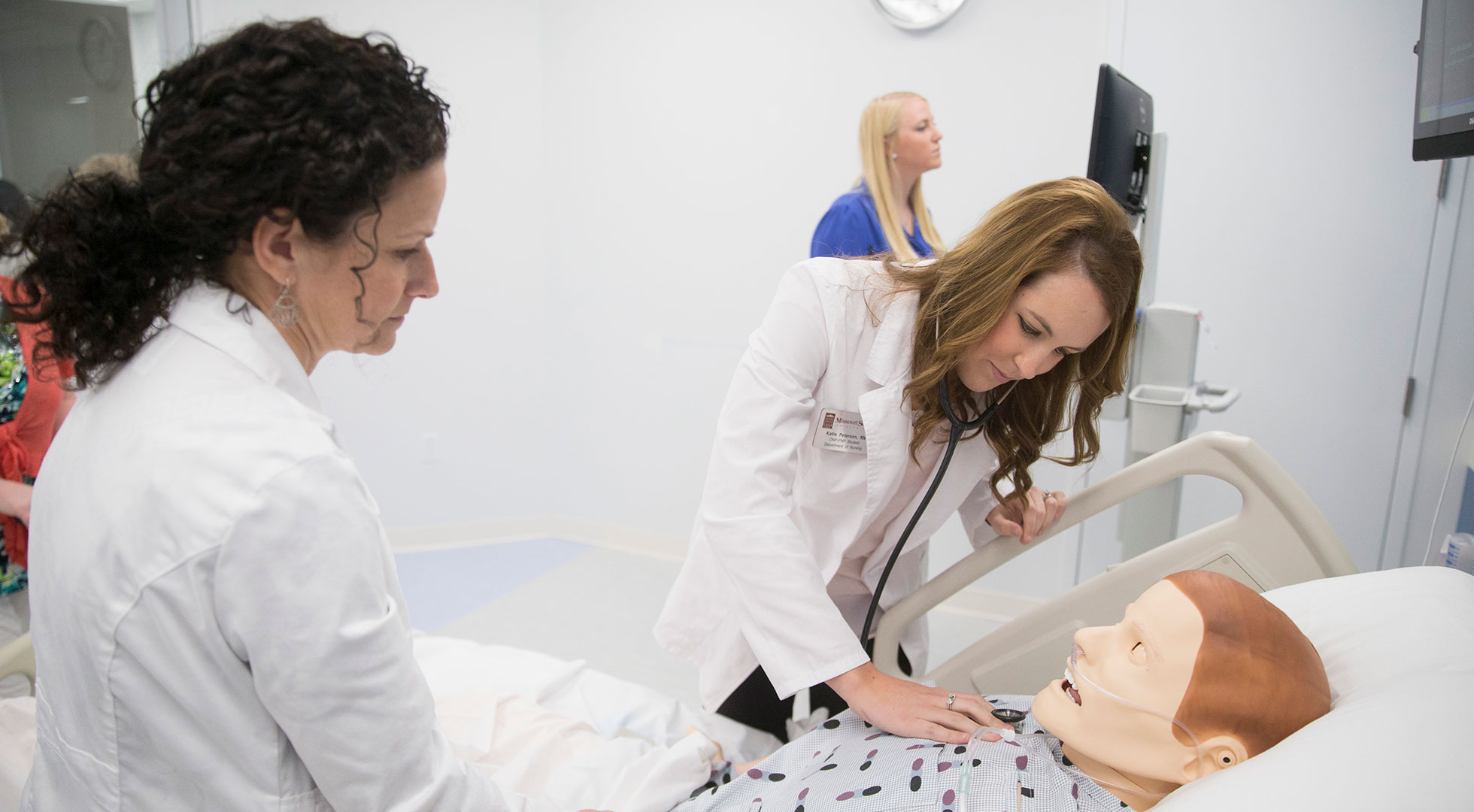 A DNP student uses a stethoscope to listen to the heartbeat of a mannequin patient, while an instructor in a white coat supervises.
