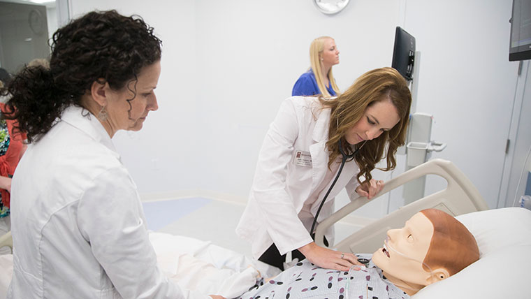 A DNP student uses a stethoscope to listen to the heartbeat of a mannequin patient, while an instructor in a white coat supervises.