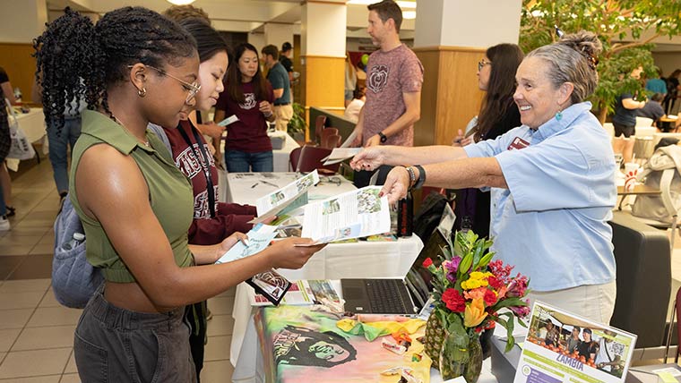 Two students receive brochures from a faculty member during an Education Abroad fair in the Plaster Student Union.