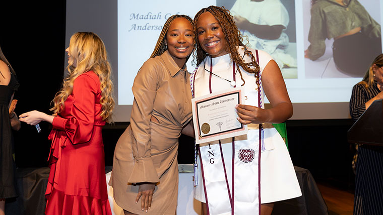 A nursing faculty member and a student pose for a photo at the pinning ceremony. The student holds a certificate for her contributions to the Student Nurse Organization.