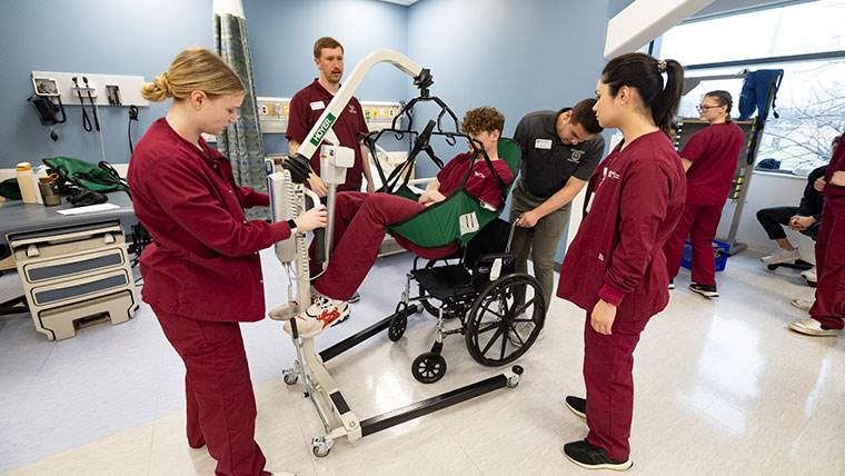 Nursing students and a physical therapy instructor transfer a student from a Hoyler lift sling into a wheelchair.