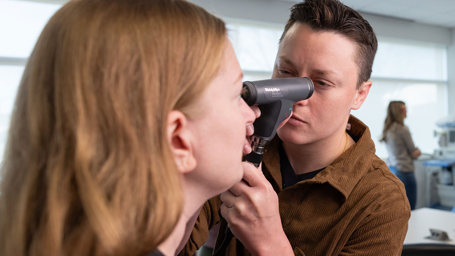 A graduate nursing student uses a tool to look into a classmate's eye.