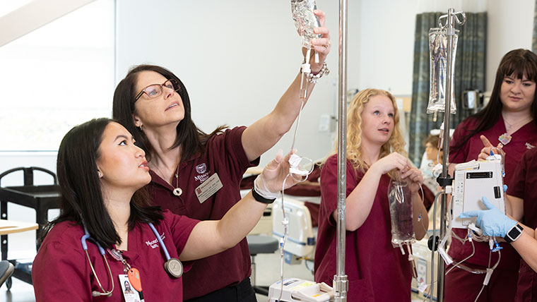 Dr. Missy Penkalski, an associate professor, shows a nursing student how to monitor the level of the fluid in an IV bag. Other nursing students are checking IV bags in the background.