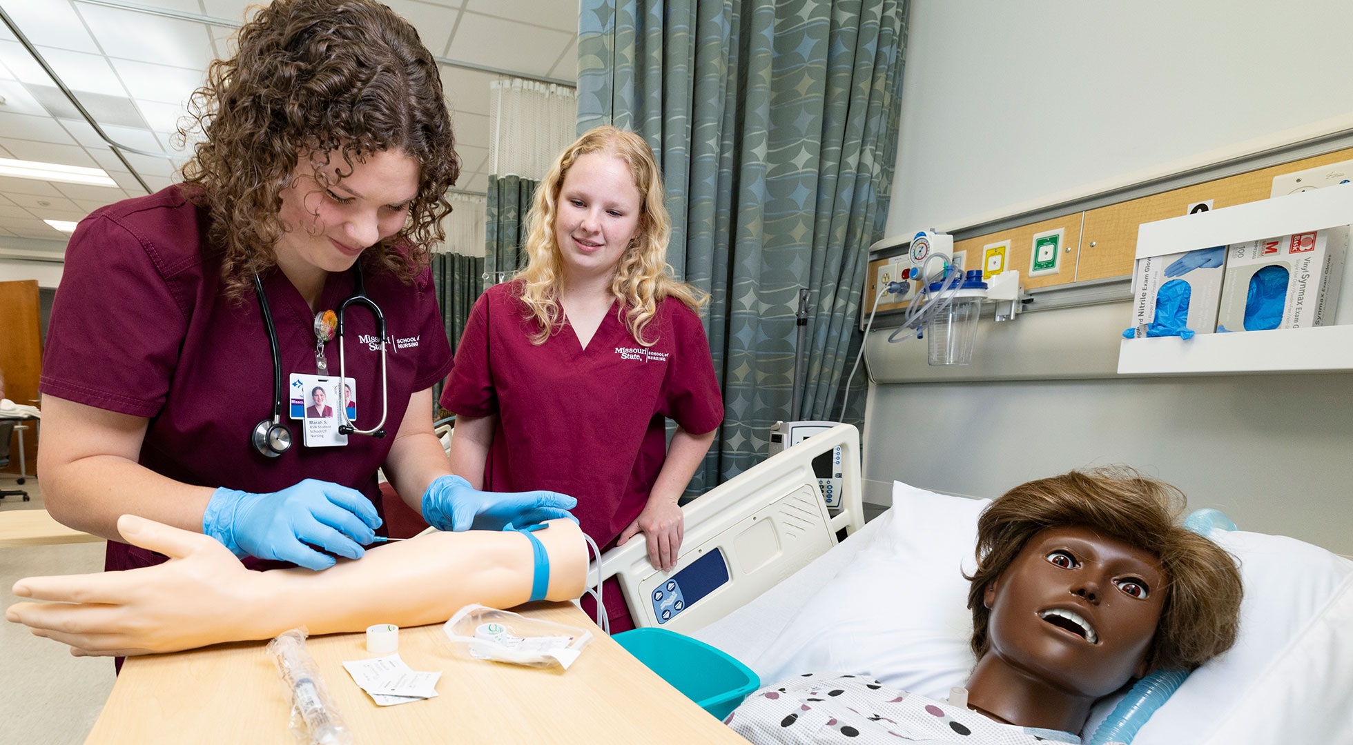 A nursing student practices administering a needle into a mannequin arm while another student observes.