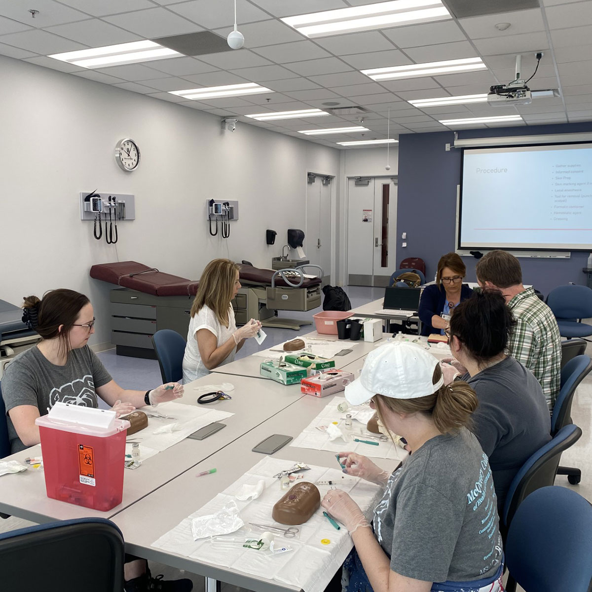 Five nursing student practice stitching up a wound, as demonstrated by their professor in a classroom.