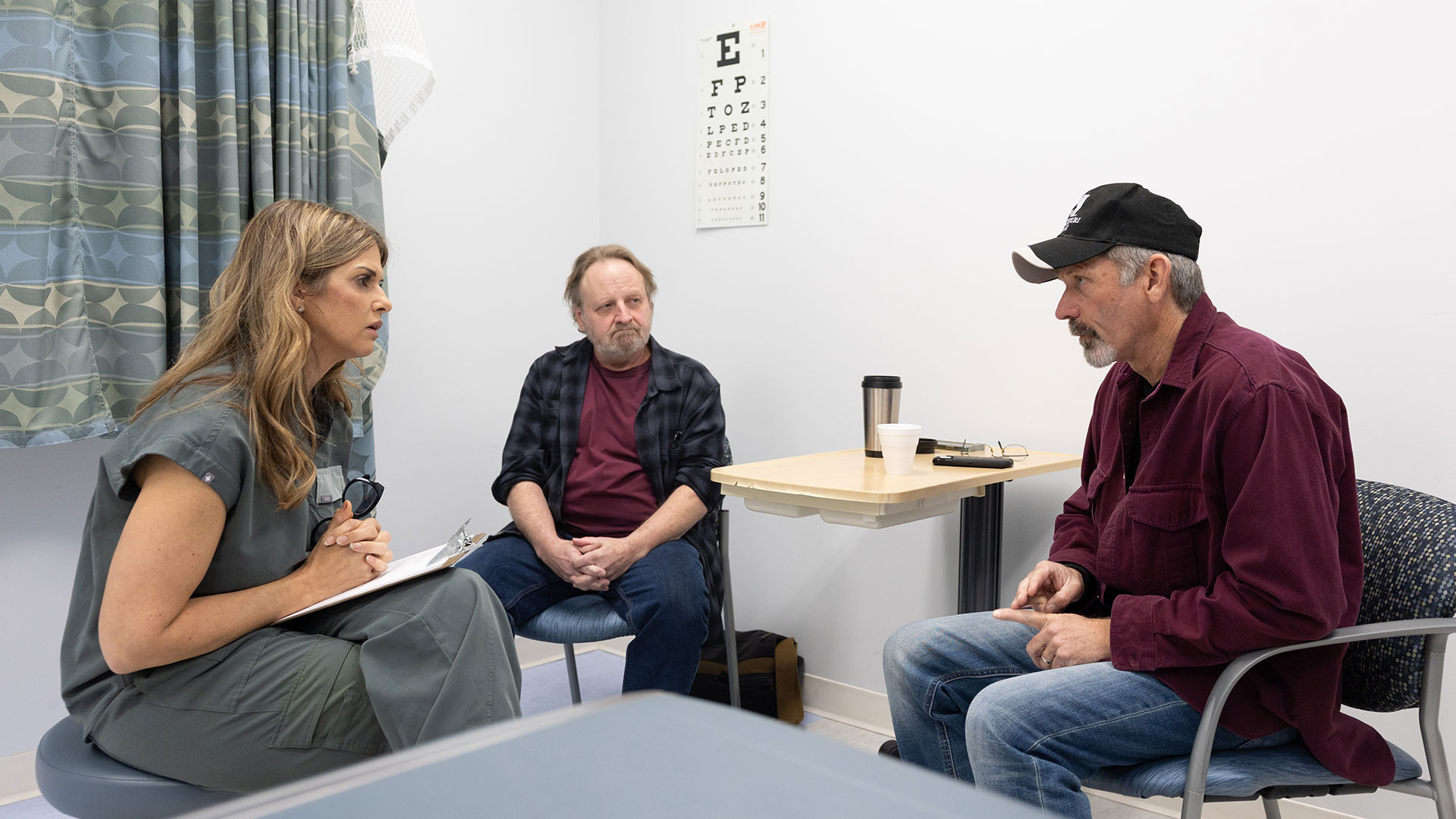 A nursing graduate student clutches a clipboard while listening to two patients.