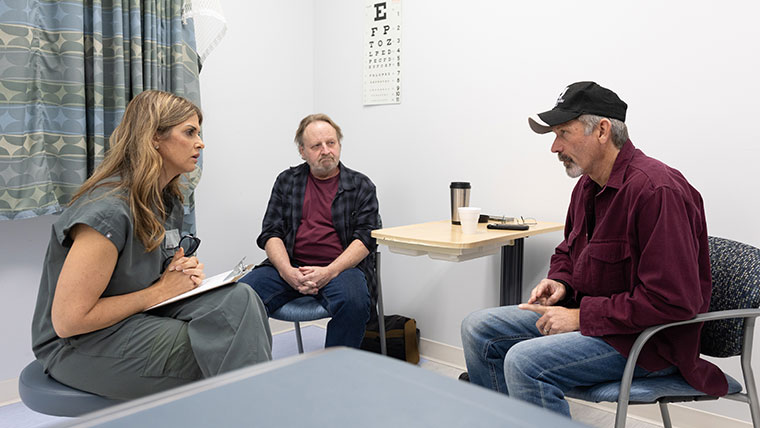 A nursing graduate student clutches a clipboard while listening to two patients.