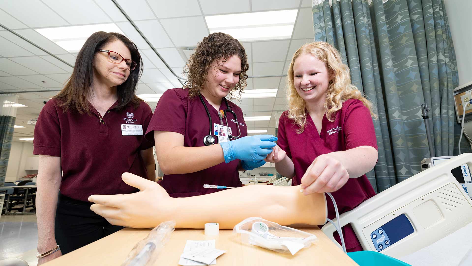 Two nursing students practice IV insertions as a faculty member supervises them.