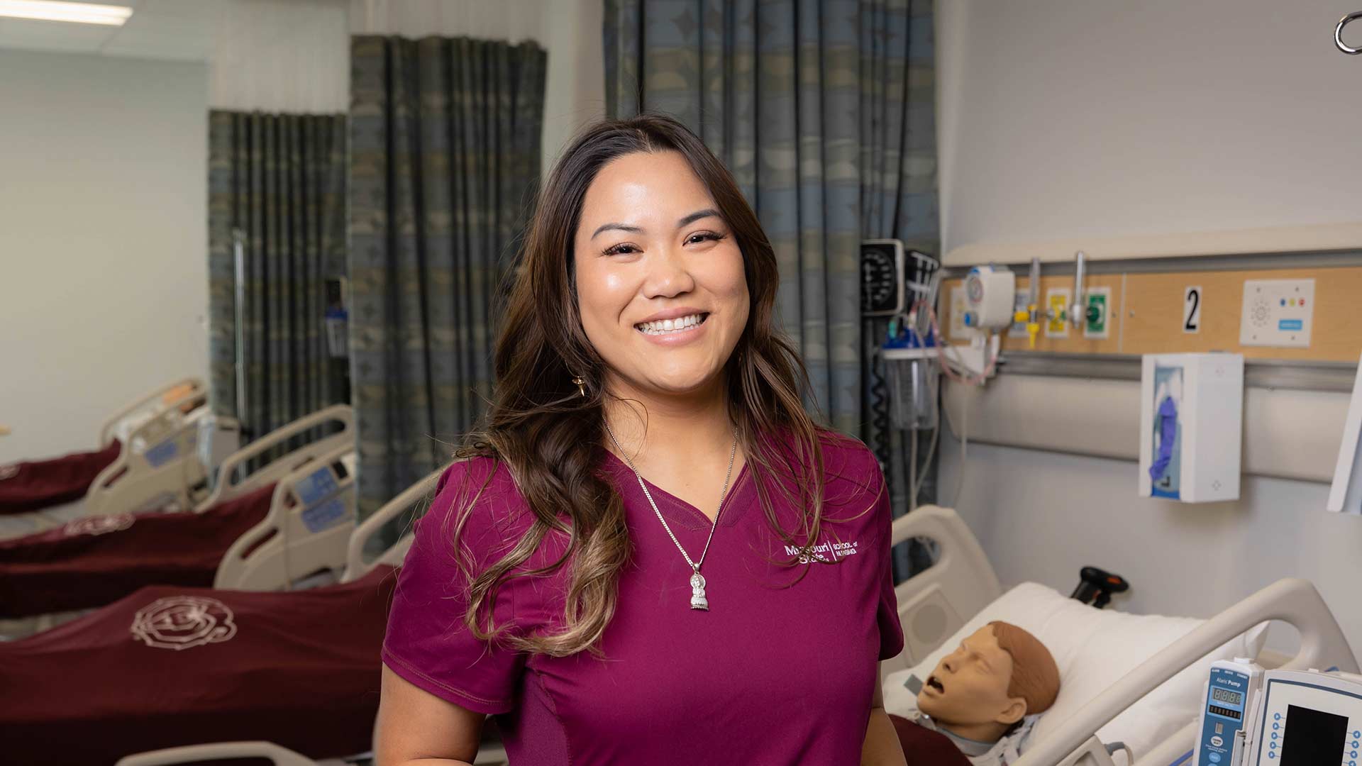 A nursing student in maroon scrubs smiles while posing in the simulation lab.
