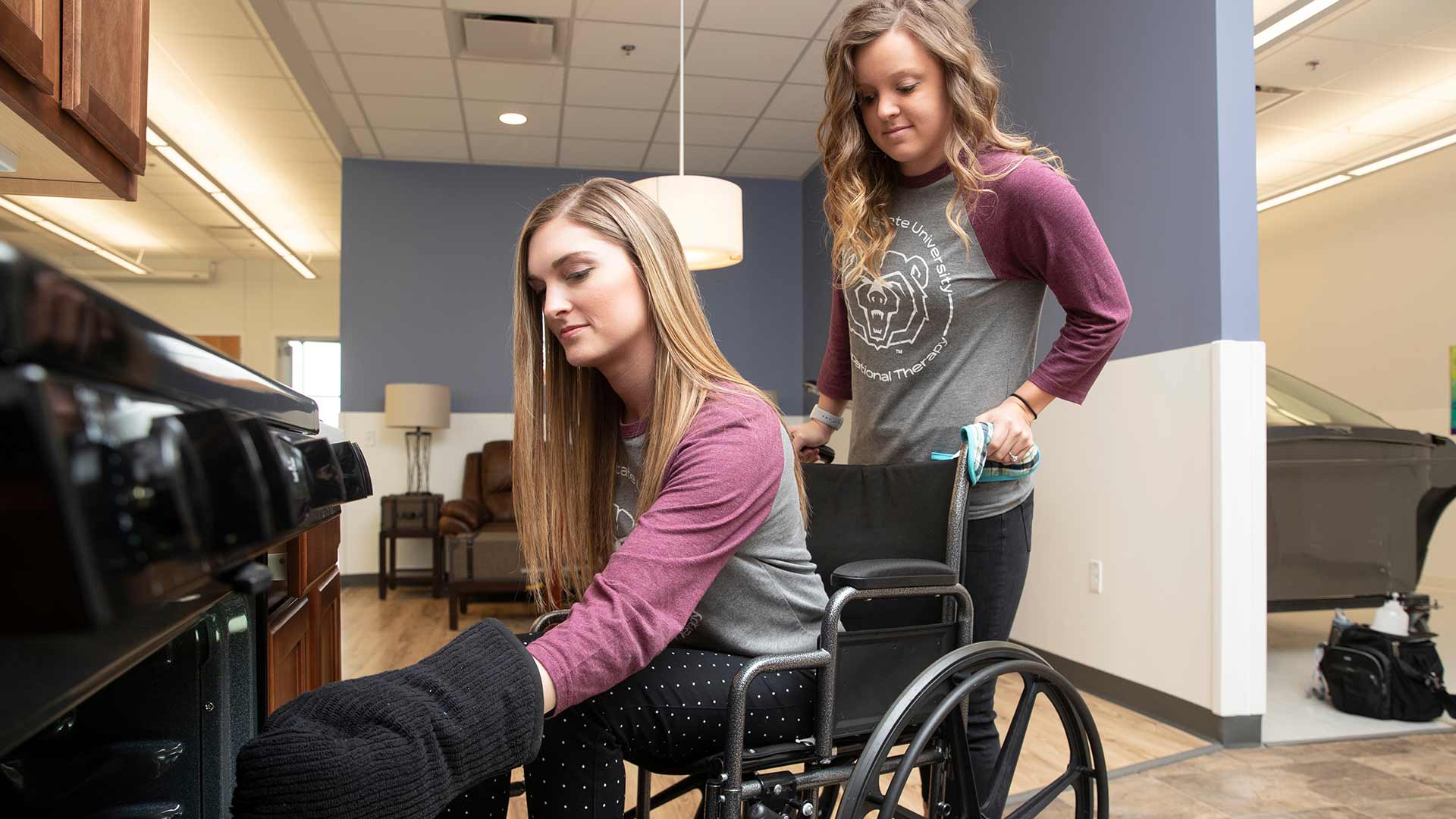 Occupational therapy student in a wheelchair using kitchen equipment while another student assists her.