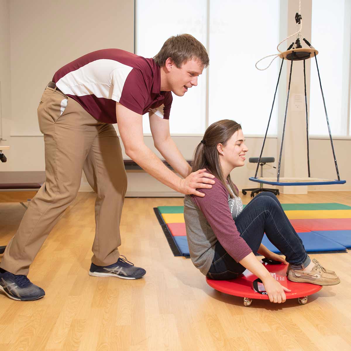Two occupational therapy students having fun in a lab. One student is pushing another sitting who is sitting down on a circular roller.