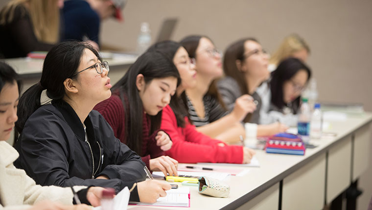 A row of accounting students seated in a lecture hall study the board while taking notes.