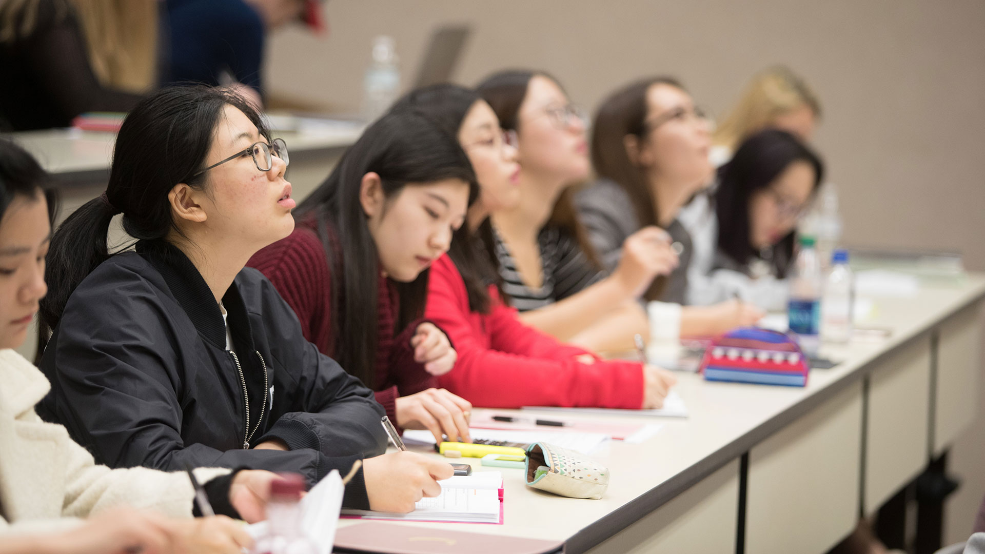 A row of accounting students seated in a lecture hall study the board while taking notes.