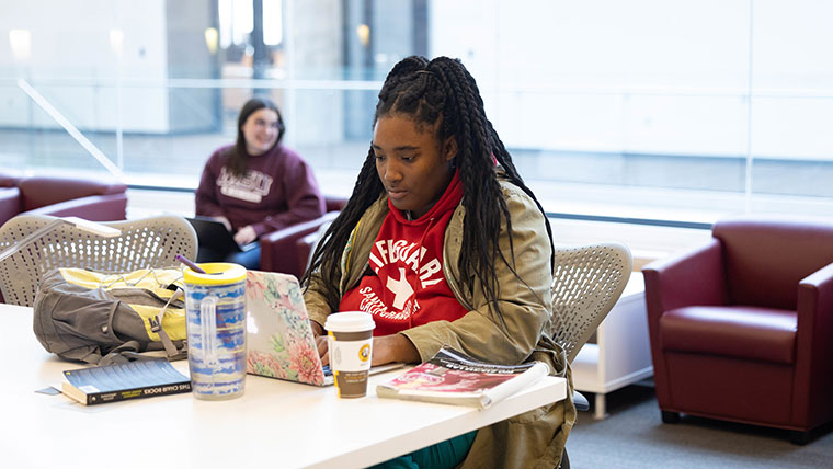 A student is seated at a table and is focused on typing on her laptop.