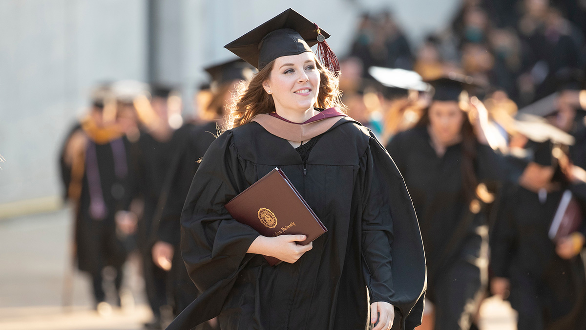 Woman in cap and gown walks with diploma.