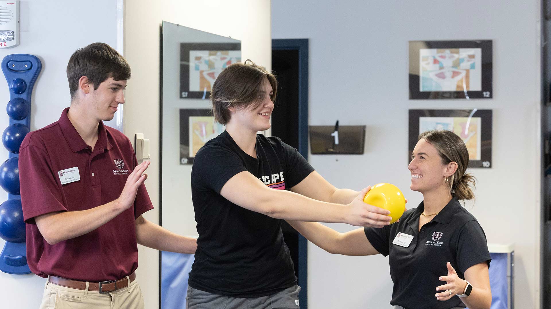Two Missouri State physical therapy students guide a teenager through a rehab activity. The teenager is moving a medicine ball from side to side.