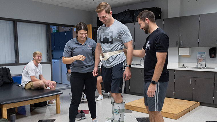 Two physical therapy students helping another student walk around in prosthetic footwear.