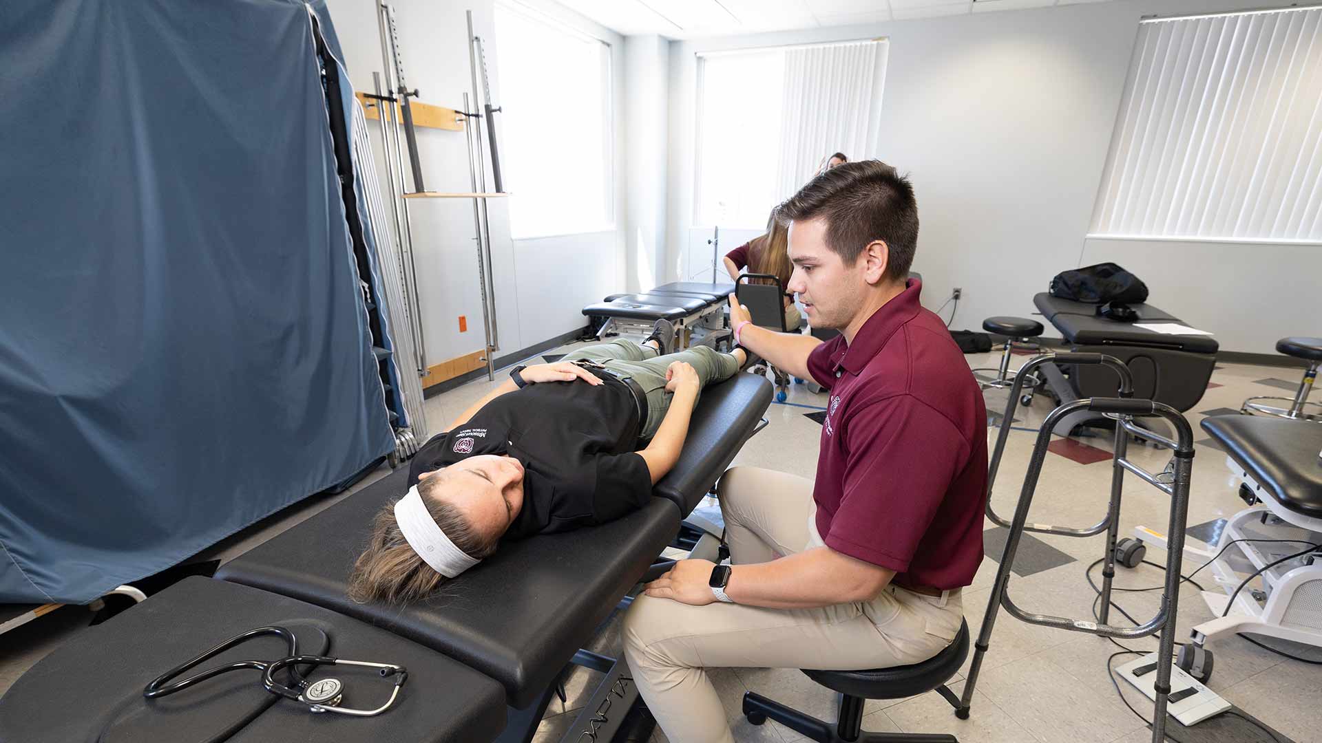 A physical therapy student seated in a chair examines another physical therapy student who is laying on a table.