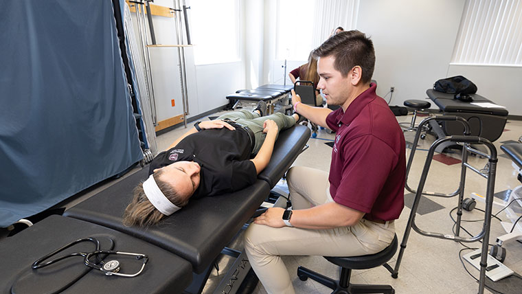 A physical therapy student seated in a chair examines another physical therapy student who is laying on a table.