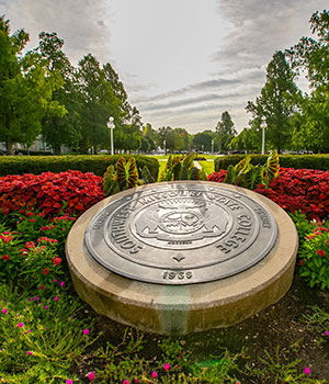 Bright sun behind clouds shines on the Missouri State official seal surrounded by bright flowers.