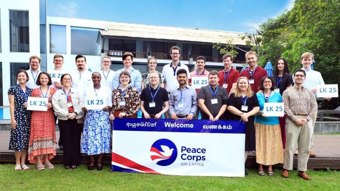 A group of people stand behind a Peace Corps Sri Lanka banner - several hold signs saying LK 25