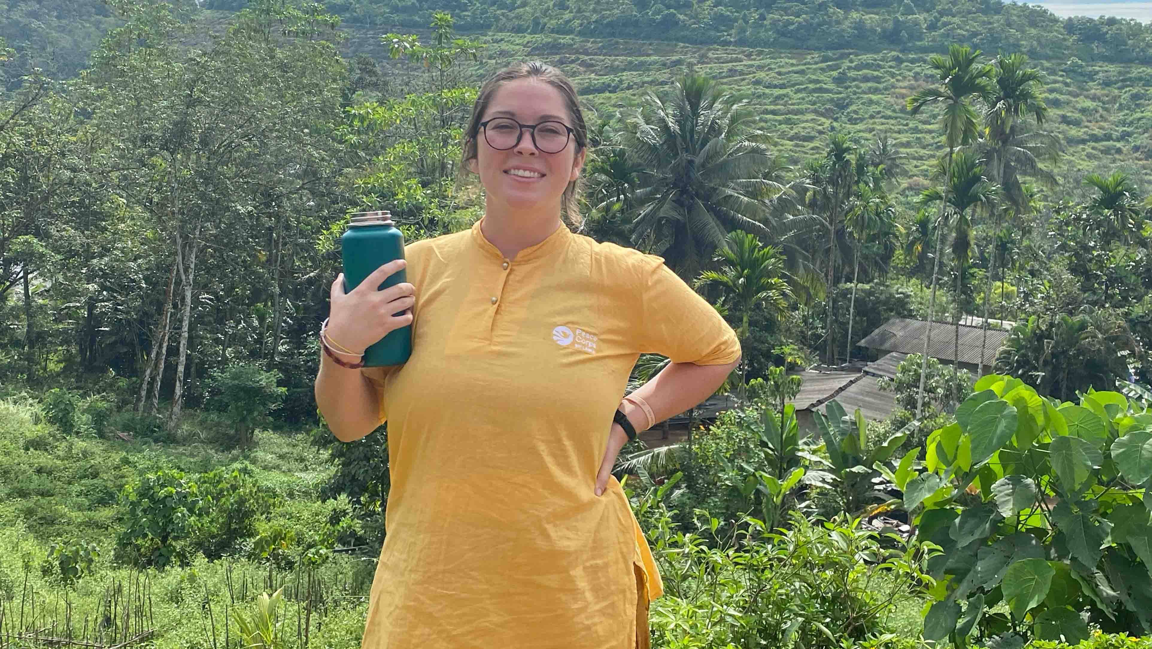 A Peace Corps volunteer smiles into the camera with lots of green vegetation behind her