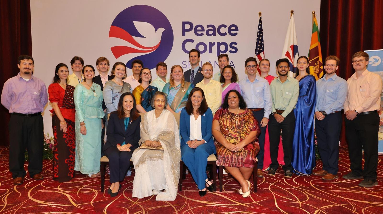 A group of people in dressy clothes in front of a Peace Corp banner and three flags
