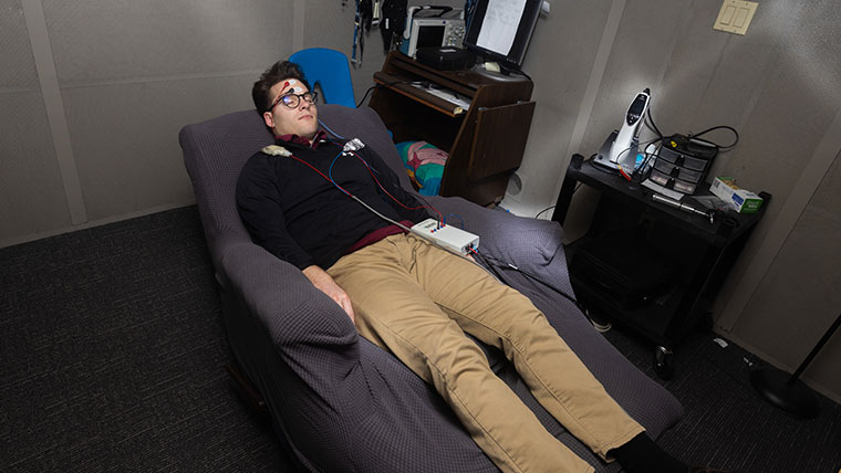 A student lays on a recliner while hooked up to testing equipment in an audiology lab.