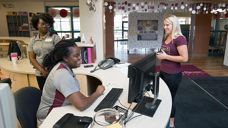 Two resident assistants - one sitting and one standing behind a desk in a dorm on the Missouri State University campus. 