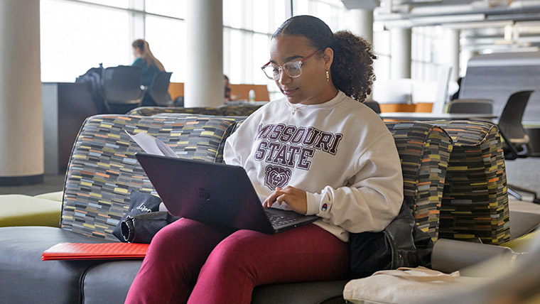 Missouri State student wearing BearWear seated within the library with laptop. 