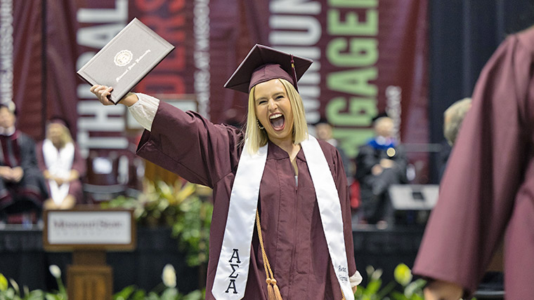 Smiling Missouri State graduate in cap and gown holding their diploma cover. 