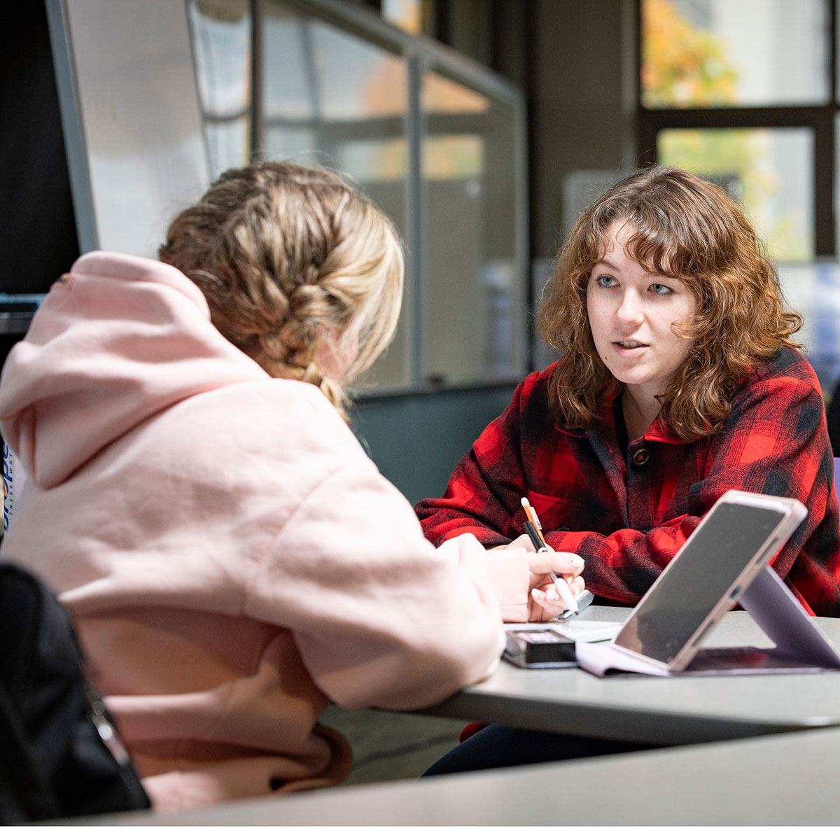 Two students have a discussion in Meyer Library. One student is listening while writing in a notebook, with her iPad at her side.