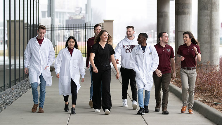 Graduate science students walk along the sidewalk. Some students are in lab coats.