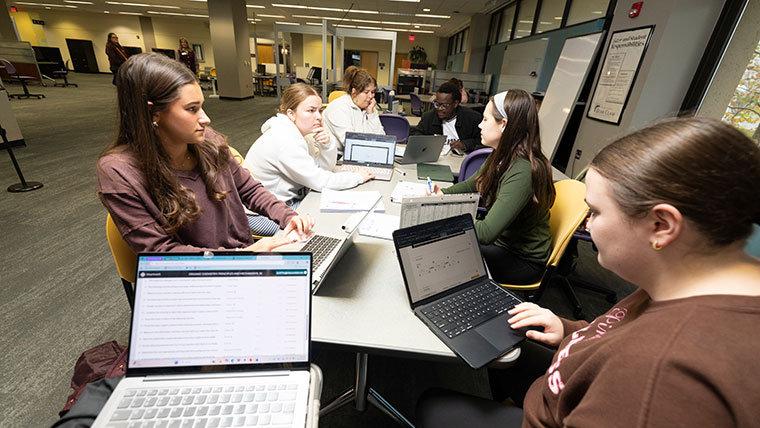 A study group of seven students sit at a table with their laptops and notebooks in the science area of the Bear CLAW.