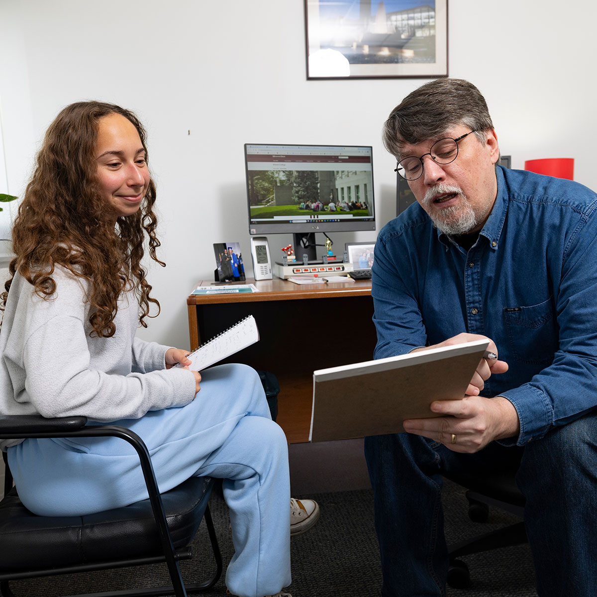An advisor shows a student an academic plan in a notebook during an advisement meeting in his office.