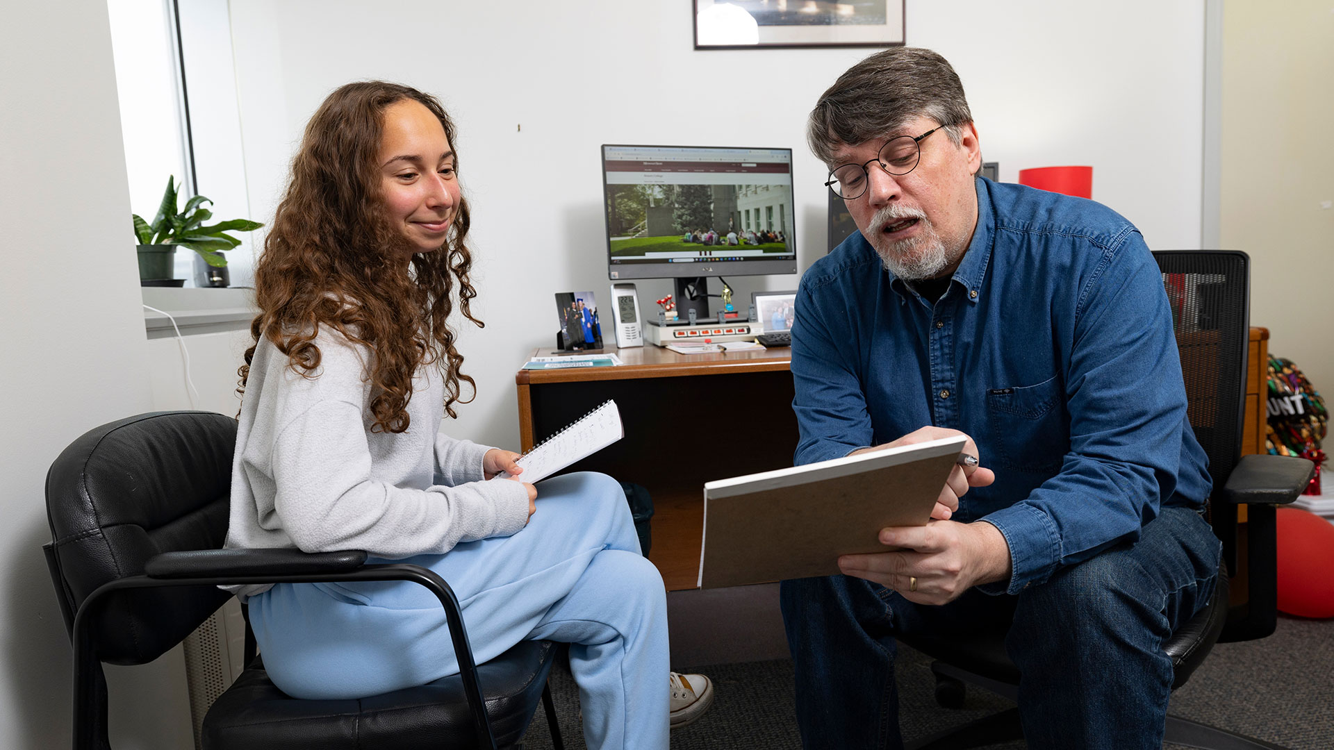 An advisor shows a student an academic plan in a notebook during an advisement meeting in his office.